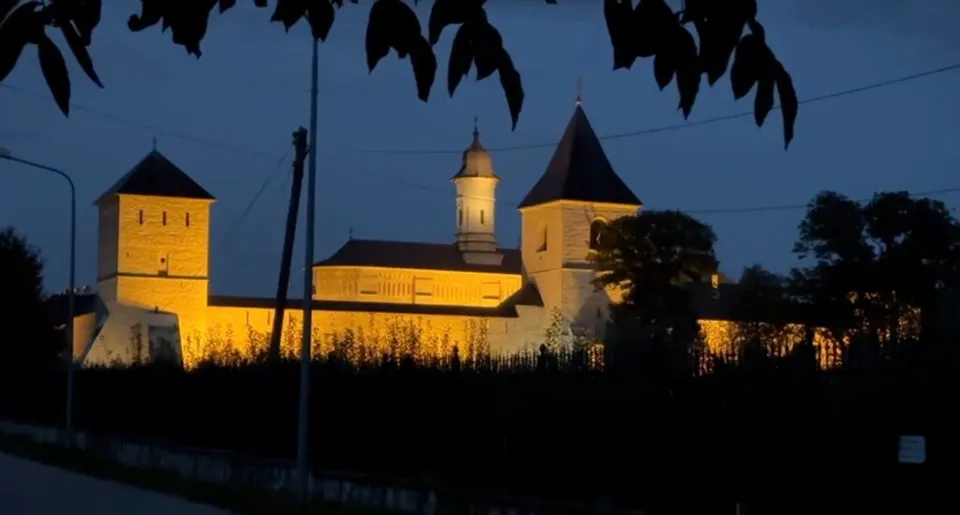 O clădire veche cu turnuri este iluminată nocturn, fiind înconjurată de vegetație sub un cer închis la culoare.