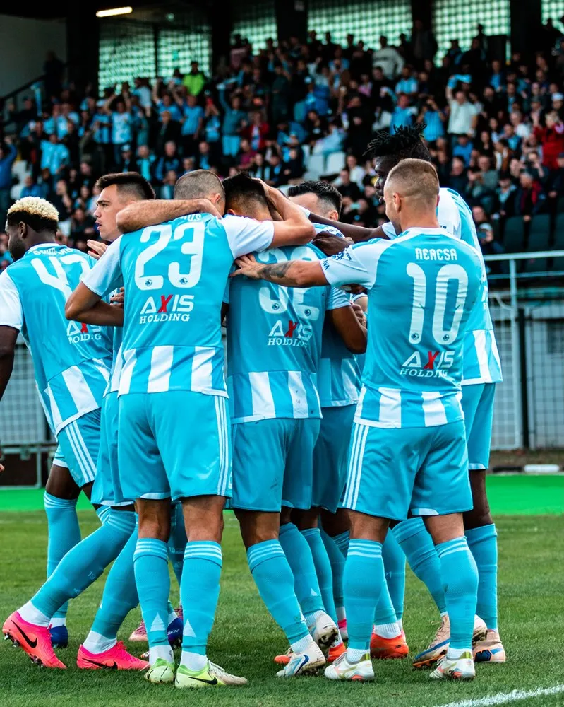 Un grup de fotbaliști în echipament albastru și alb se îmbrățișează pe un teren de fotbal în fața unor tribune cu spectatori.