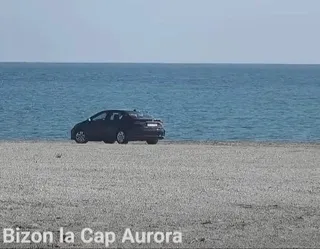 Un autoturism negru este staționat pe o suprafață nisipoasă în fața unei mări sub un cer senin.