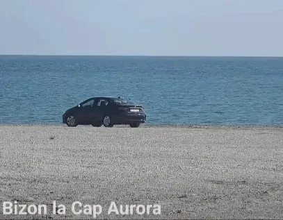 Un autoturism negru este staționat pe o suprafață nisipoasă în fața unei mări sub un cer senin.