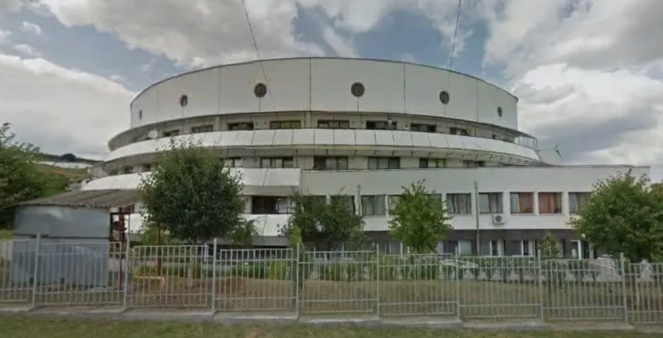 O clădire albă, circulară, cu mai multe etaje, situată în spatele unui gard metalic și unor copaci verzi sub un cer înnorat.