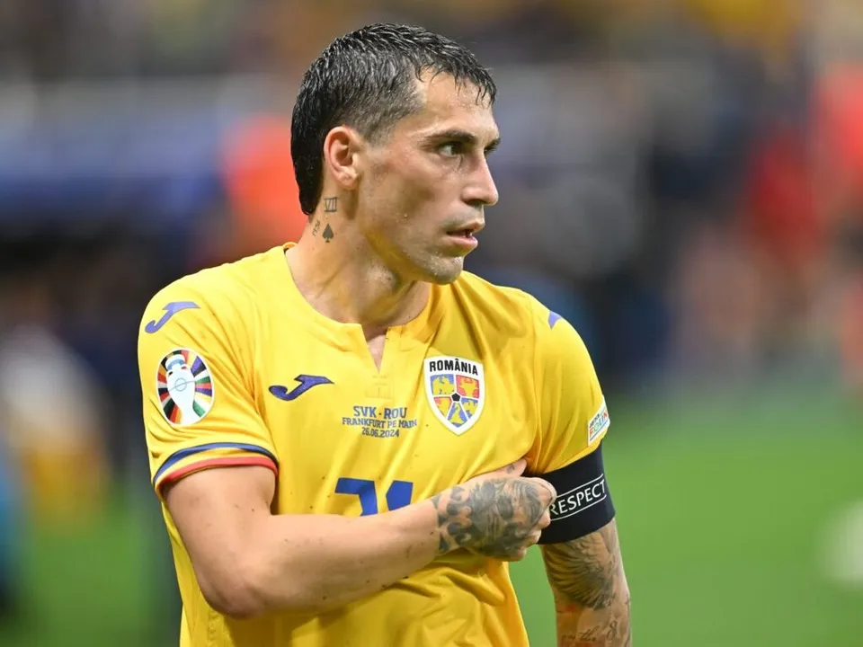 Un bărbat cu părul scurt și tatuaje pe gât și braț poartă un tricou galben cu embleme și un brățar negru pe braț.