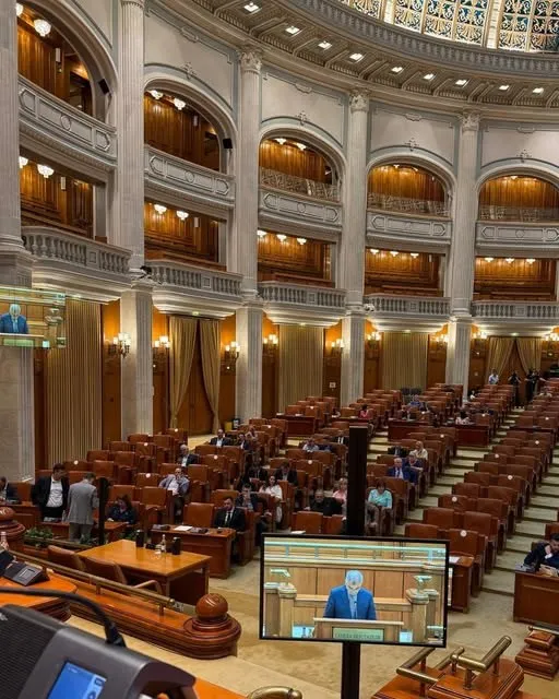 O sală de conferințe mare cu rânduri de scaune din lemn, balcone înarcuite și un monitor afișând un bărbat la un pupitru.