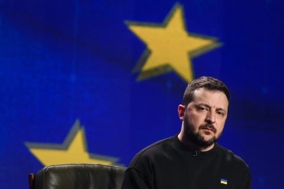 Un bărbat cu barbă și păr scurt, purtând un tricou negru, stă în fața unui fundal albastru cu stele galbene.
