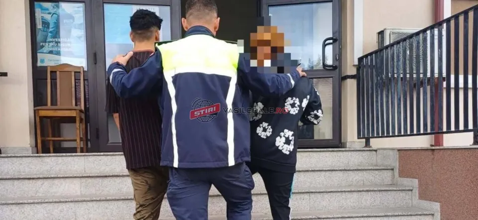Un bărbat cu o jachetă albastră și galbenă stă cu brațele întinse pe umerii a doi tineri care merg pe scări în fața unei clădiri.