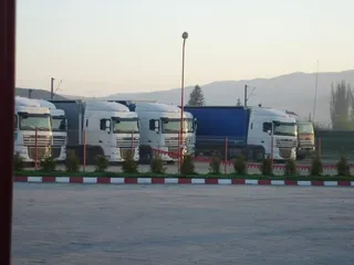 Cinci camioane parate într-o parcare lângă un gard, sub un cer luminos și dealuri în fundal.