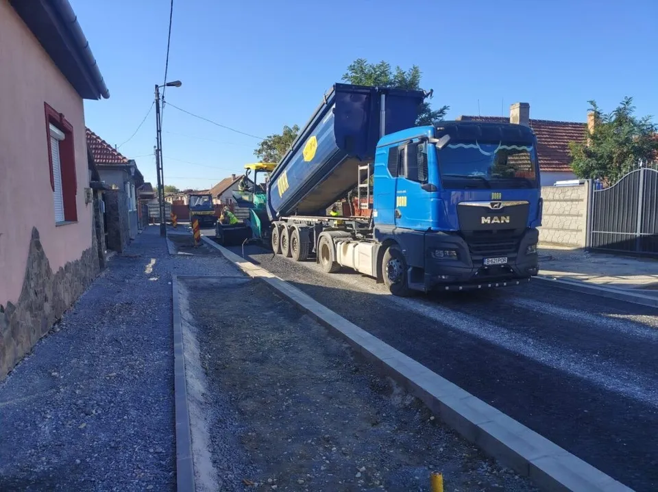 Un camion albastru cu remorcă basculantă ridicată stă pe un drum cu asfalt nou lângă o bordură de beton și case.