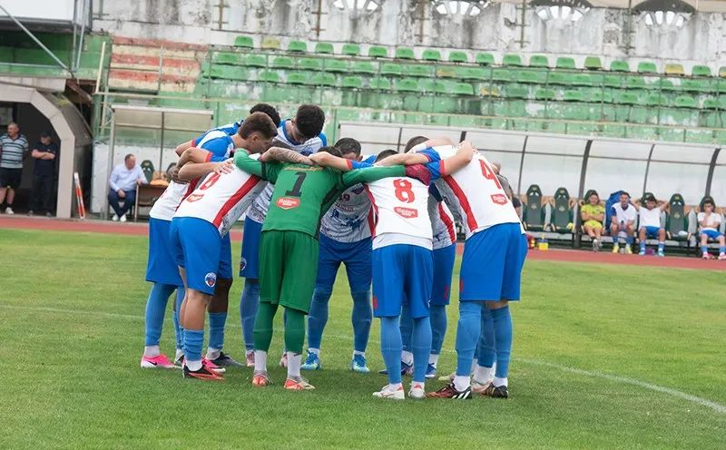 O echipă de fotbaliști în echipament sportiv albastru și roșu stă într-un cerc pe un teren de gazon verde.