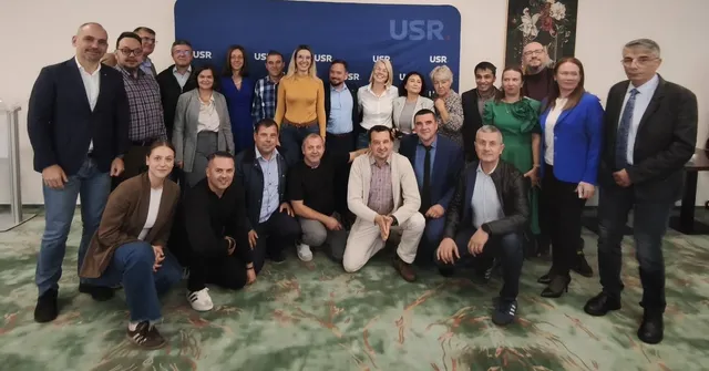 Un grup de oameni stă poziționat în două rânduri în fața unui panou albastru cu inscripții albe, într-o încăpere cu podea cu modele verzi.