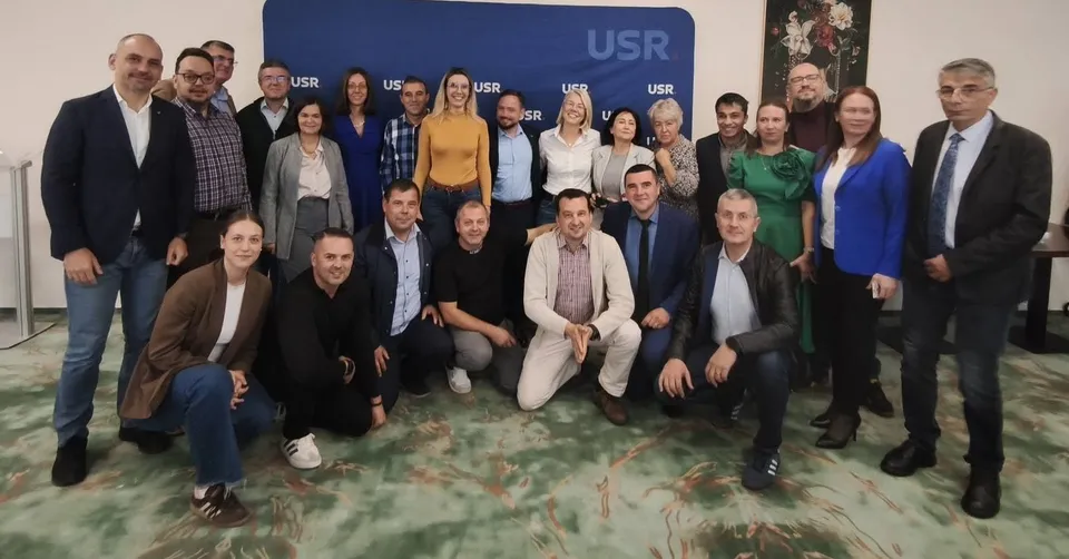 Un grup de oameni stă poziționat în două rânduri în fața unui panou albastru cu inscripții albe, într-o încăpere cu podea cu modele verzi.