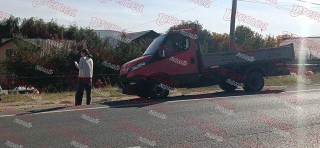Un camion roșu este oprit pe marginea unui drum lângă o persoană care poartă o cămașă albă și pantaloni închiși.