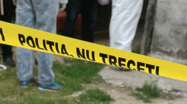 O bandă galbenă cu text negru este întinsă pe un teren cu iarbă și pământ, în fața unor persoane care stau în picioare.