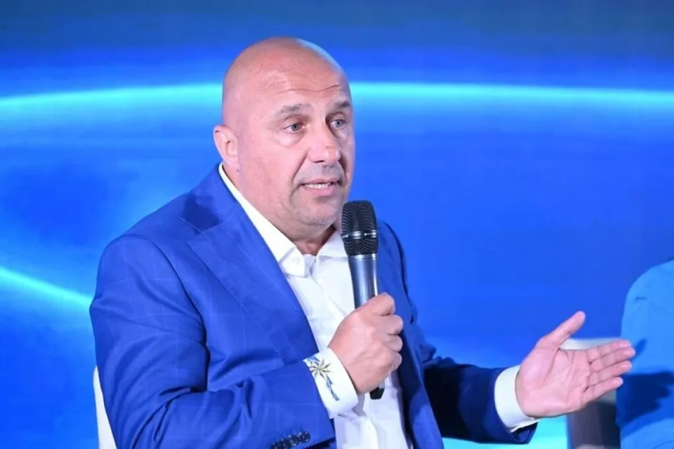 Un bărbat cu capul chel, purtând un sacou albastru și cămașă albă, ține un microfon negru în mână.