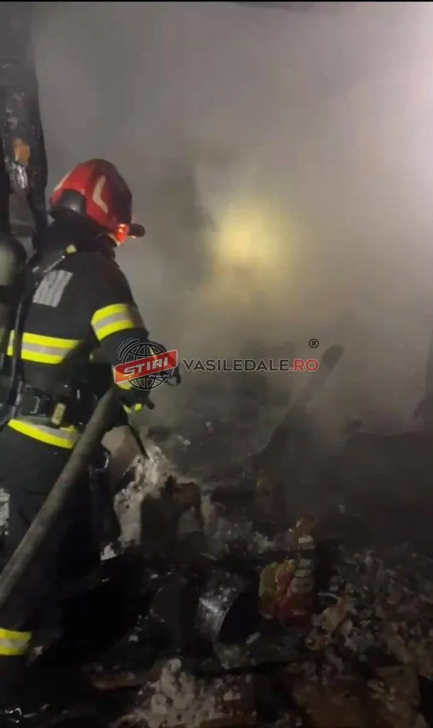 Un pompier cu echipament de protecție și cască este orientat spre o zonă plină de fum și resturi arse.