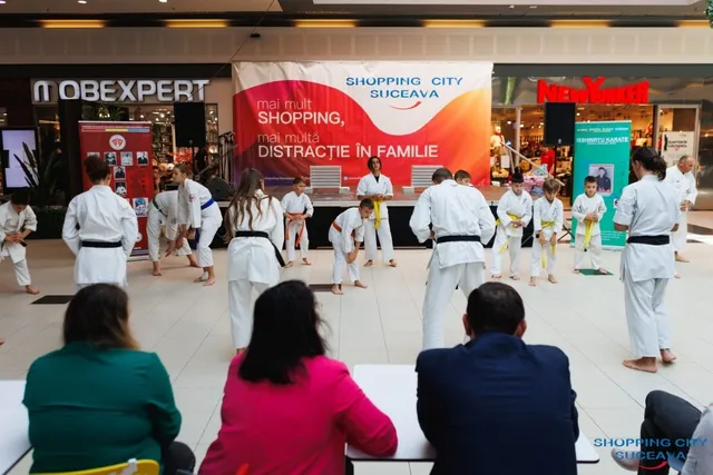 Mai mulți copii îmbrăcați în uniforme albe de karate execută mișcări pe un podea de mall, în fața unor spectatori așezați.