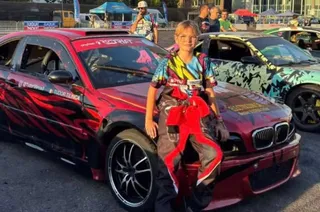 Un adolescent îmbrăcat într-un costum colorat stă sprijinit de capota unei mașini roșii decorate cu modele negre, lângă o altă mașină decorată.