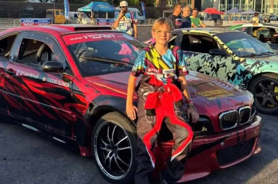 Un adolescent îmbrăcat într-un costum colorat stă sprijinit de capota unei mașini roșii decorate cu modele negre, lângă o altă mașină decorată.