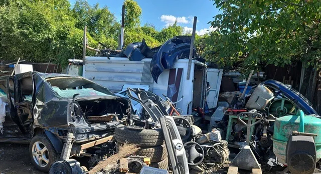 Un grup de piese auto și resturi metalice sunt depozitate într-un spațiu exterior lângă un vehicul alb și un autoturism negru parțial demontat.
