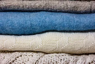 Mai multe haine din material textil tricotat, de culori gri, albastru și alb, sunt așezate unele peste altele.