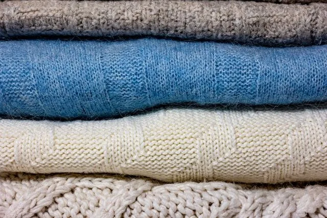 Mai multe haine din material textil tricotat, de culori gri, albastru și alb, sunt așezate unele peste altele.