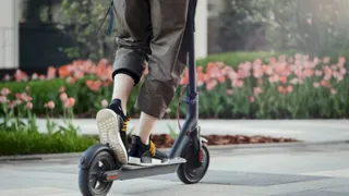 O persoană poartă pantaloni de culoare maro și încălțăminte neagră cu talpă albă în timp ce stă pe o trotinetă electrică neagră pe un drum pavat.