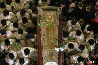 Oamenii în haine de culoarea nisipului și căciuli negre stau în jurul unui obiect lung, dreptunghiular, decorat cu o figură aurie.