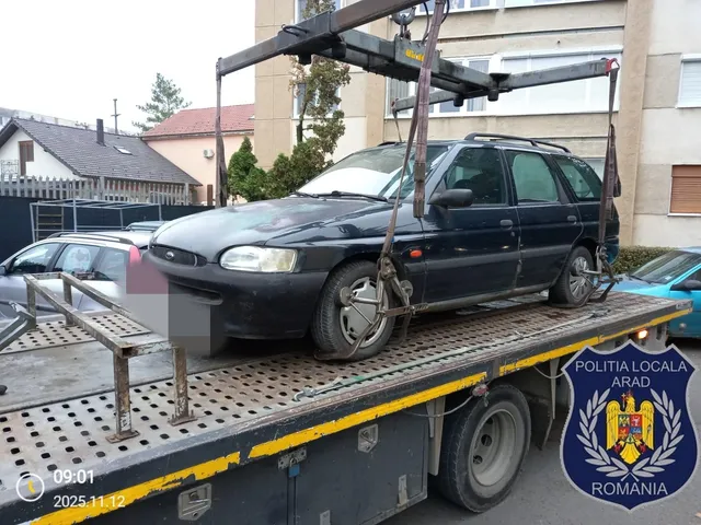 Un autoturism de culoare închisă este transportat pe platforma unei mașini de remorcare cu ajutorul unor curele de fixare.