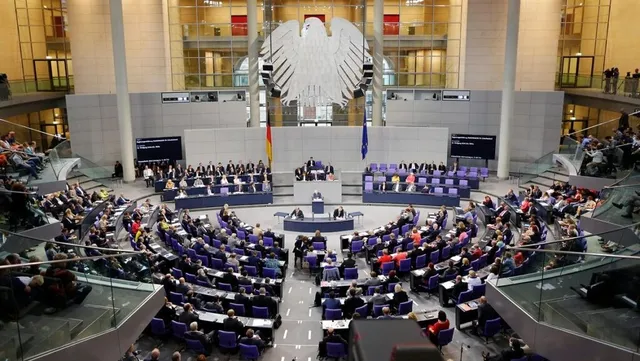 O mulțime de oameni stă așezată la mese în semicerc într-o sală mare cu pereți înalți și un simbol al unei păsări mari pe perete.