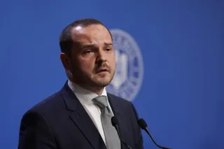 Un bărbat cu barbă, purtând sacou închis la culoare, cămașă albă și cravată gri, stă în fața unui fundal albastru cu un emblemă circulară.