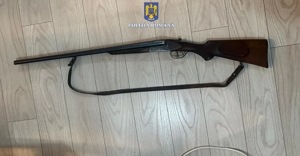 O armă de foc cu țevă lungă și trăsătură de lemn este întinsă pe un podea de lemn deschis la culoare.