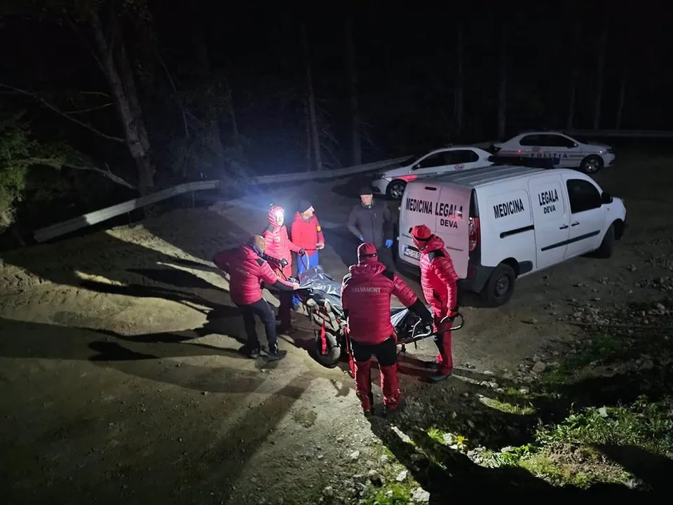 Patru persoane în haine roșii transportă o targă pe un teren accidentat lângă un van alb în timpul nopții.