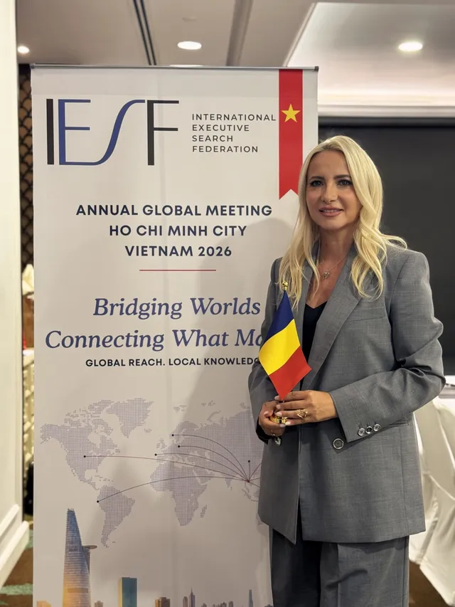 O femeie cu părul blond, purtând un costum gri, stă lângă un panou informativ și ține un mic steag românesc.