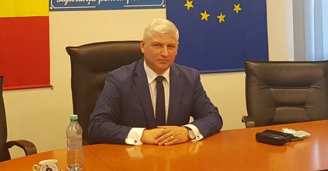Un bărbat cu părul alb, purtând costum închis la culoare și cravată, stă la o masă de lemn în fața unor steaguri.