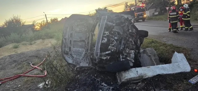 Un autoturism negru este răsturnat pe o parte într-un șanț, lângă un parapet de beton, în timp ce personal de intervenție stă pe drum.