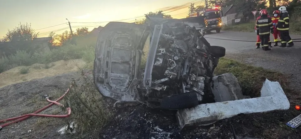 Un autoturism negru este răsturnat pe o parte într-un șanț, lângă un parapet de beton, în timp ce personal de intervenție stă pe drum.