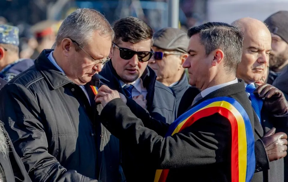 Un grup de bărbați în vârstă de medie și bătrâni, unul purtând o eșarfă tricoloră, se află într-o mulțime în aer liber, purtând haine de iarnă și ochelari de soare.