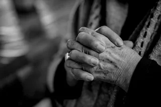 O fotografie alb-negru, cu mâinile încrețite ale unei persoane în vârstă, împreunate, cu un inel pe degetul inelar.