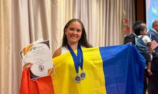 O fată zâmbește purtând două medalii și ținând un certificat și un steag tricolor în față.