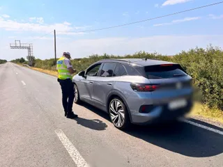 Un agent de poliție cu vest reflectorizant stă lângă o mașină gri oprită pe marginea unui drum asfaltat sub un cer senin.