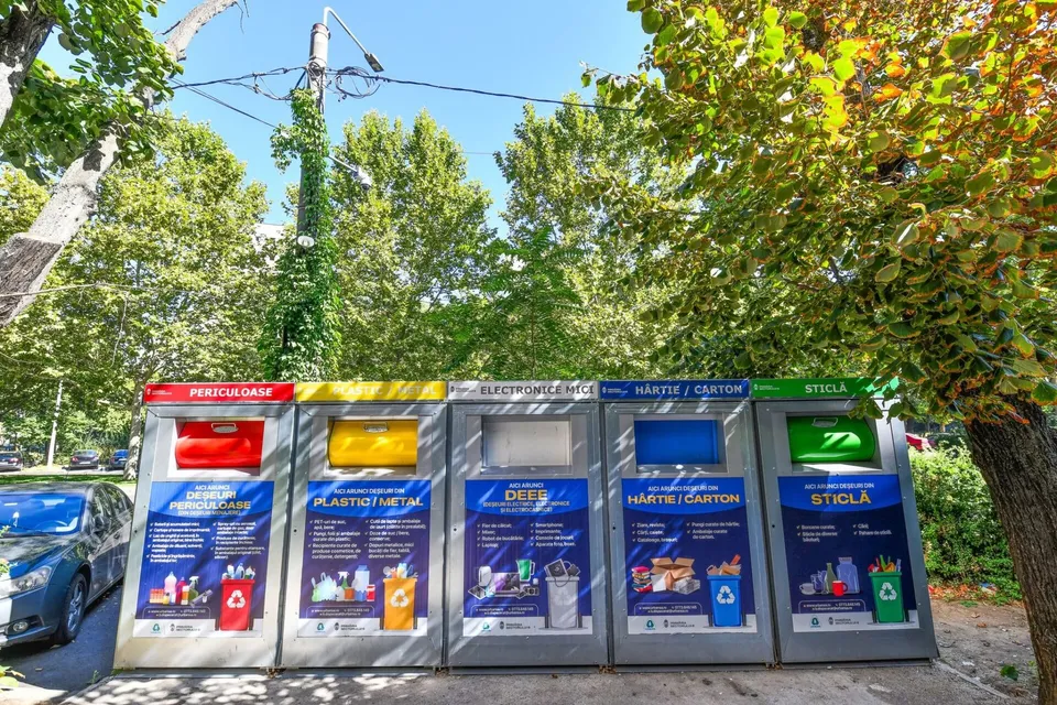 Cinci containere colorate pentru reciclare sunt aliniate pe un trotuar lângă copaci verzi sub un cer senin.