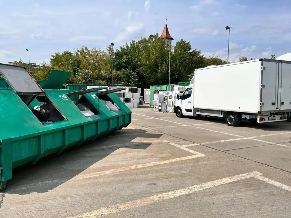 Mai multe containere verzi de colectare sunt așezate pe un teren asfaltat lângă un camion alb parcat.
