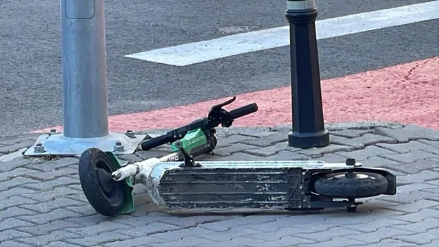 O trotinetă electrică de culoare verde și gri este întinsă pe un trotuar pavat lângă un stâlp metalic și un bolard negru.