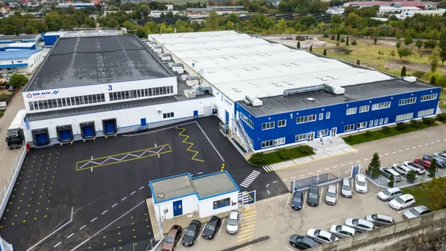 O vedere aeriană a unei clădiri industriale mari cu un acoperiș gri și albastru, parcare cu mai multe mașini și o zonă de încărcare.