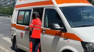 Un om într-o uniformă portocalie și albastră stă lângă o dubă albă cu dungi portocalii pe un drum asfaltat.
