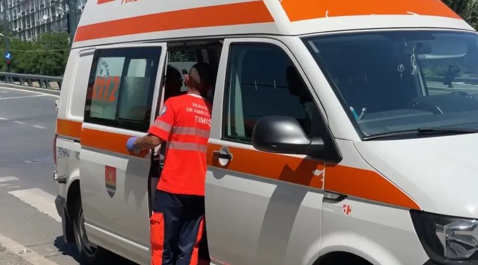 Un om într-o uniformă portocalie și albastră stă lângă o dubă albă cu dungi portocalii pe un drum asfaltat.