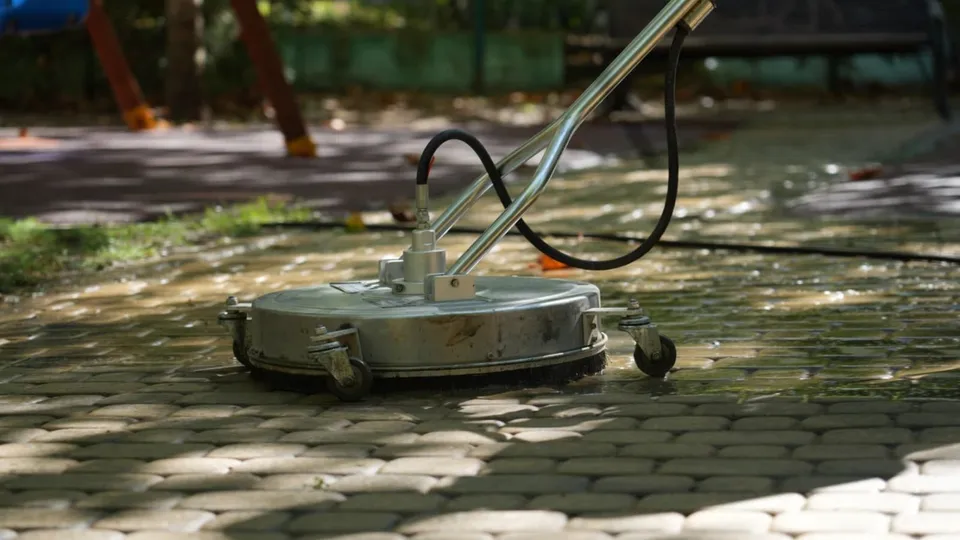 O mașină de spălat cu apă este poziționată pe un pavaj umed într-o zonă cu copaci în fundal.