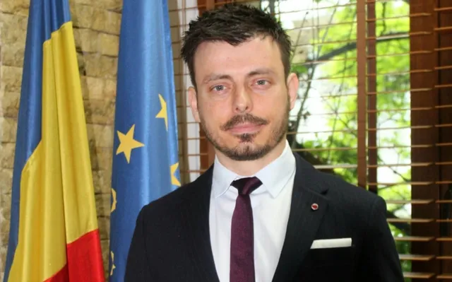 Un bărbat cu barbă și mustață, purtând sacou negru, cămașă albă și cravată purpurie, stă în fața unui steag și al unei ferestre cu jaluzele.