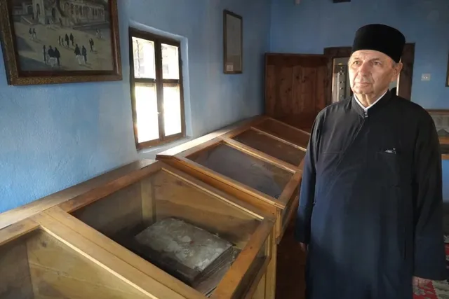 Un bărbat cu pălărie neagră și haine închise stă într-o încăpere cu pereți albaștri lângă vitrine de lemn care conțin obiecte.