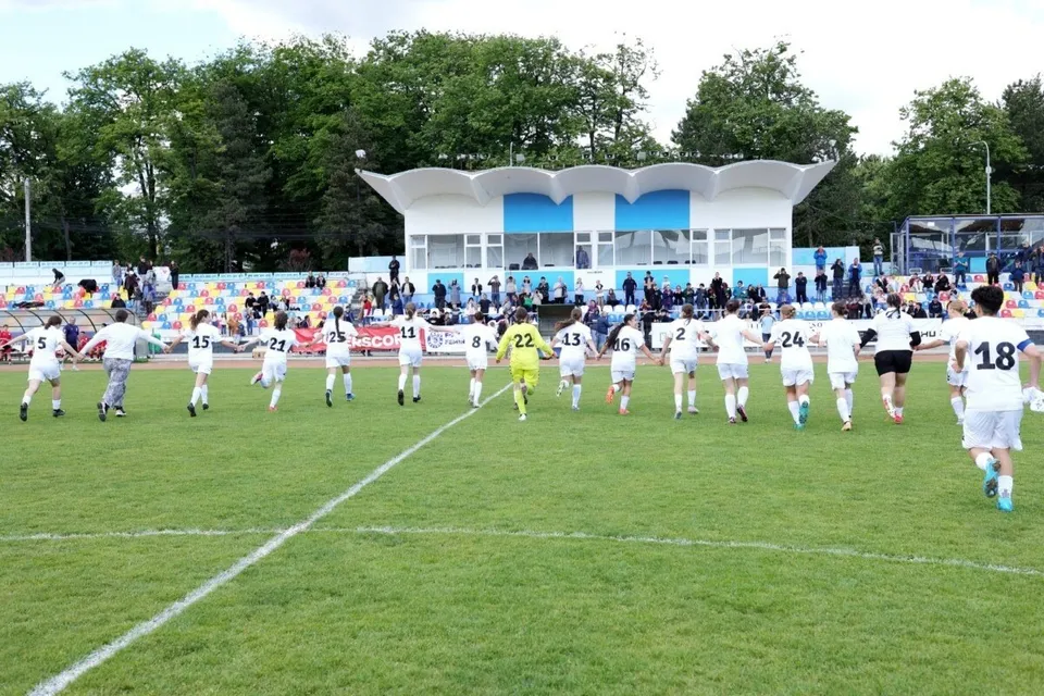 O echipă de fotbal feminin îmbrăcată în albal pornește spre centrul terenului de fotbal pe un gazon verde sub un cer înnorat.