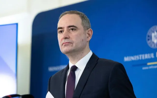 Un bărbat cu părul scurt, purtând sacou închis la culoare, cămașă albă și cravată purpurie, stă în fața unui fundal albastru cu inscripții albe.